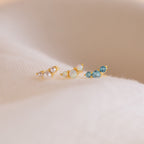 The Bella Birthstone Studs, a set of three gold stud earrings with pearls, opals, and blue gems on soft beige fabric, make a perfect personalized gift for mothers and daughters.