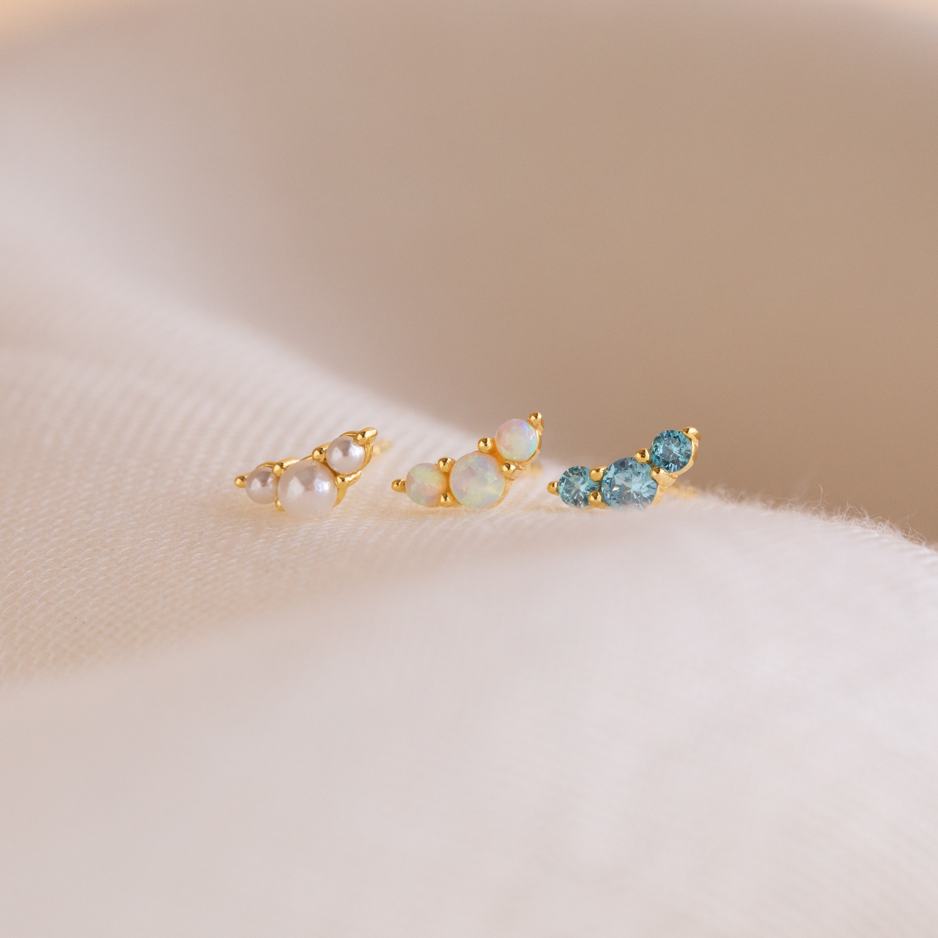 The Bella Birthstone Studs, a set of three gold stud earrings with pearls, opals, and blue gems on soft beige fabric, make a perfect personalized gift for mothers and daughters.