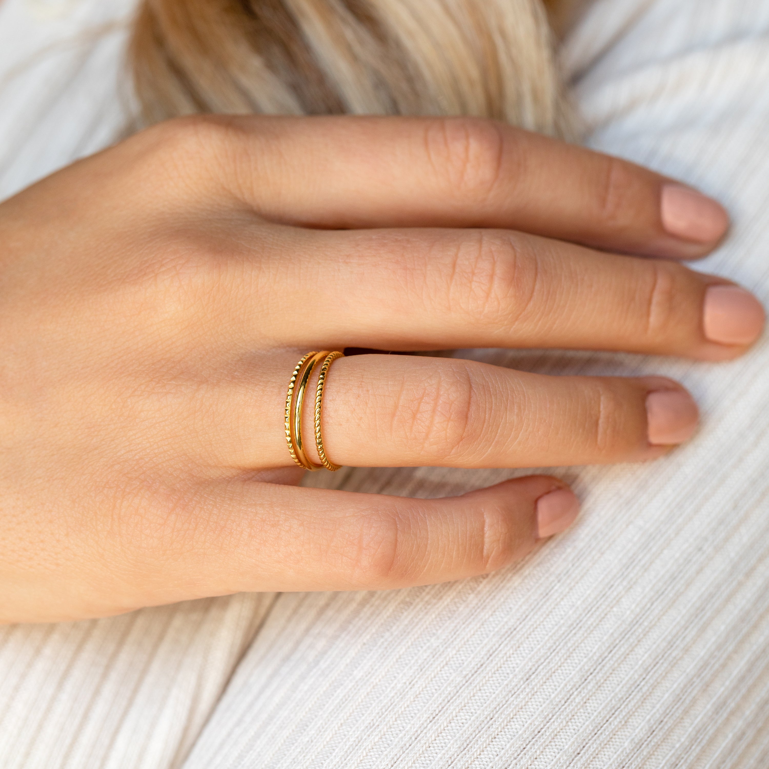 A hand with nude nail polish models the Thin Stacking Rings Set—two gold bands stacked on the ring finger, resting elegantly on white fabric.