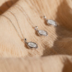 The Initial Pendant Necklace in Sterling Silver rests on textured beige fabric, softly illuminated by natural sunlight, showcasing its vintage-inspired round pendant style.