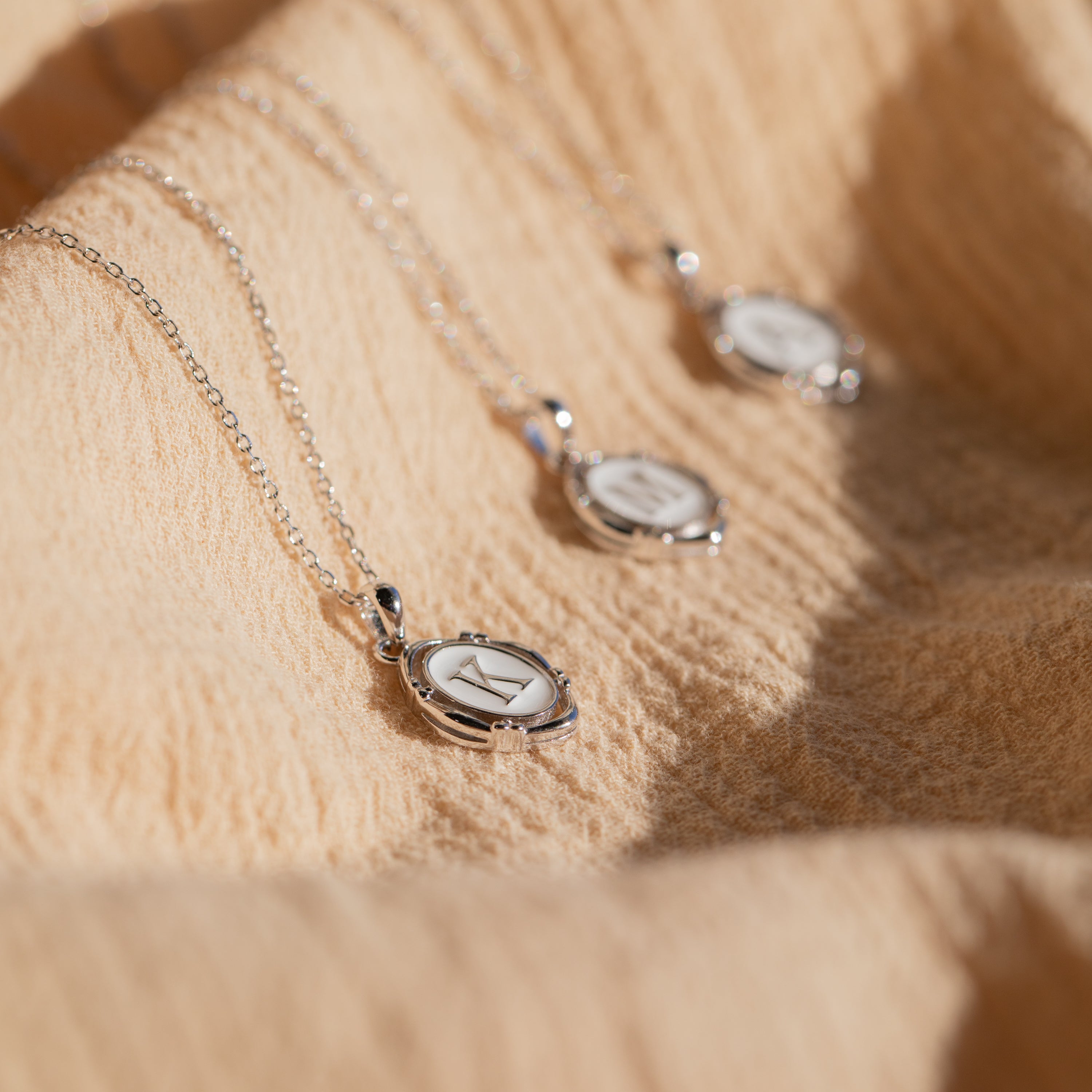 The Initial Pendant Necklace in Sterling Silver rests on textured beige fabric, softly illuminated by natural sunlight, showcasing its vintage-inspired round pendant style.