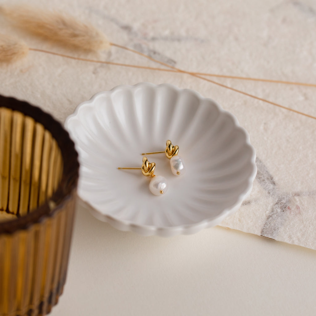 Knot Pearl Drop Earrings displayed in a white scalloped dish, accompanied by a brown glass and dried grass.