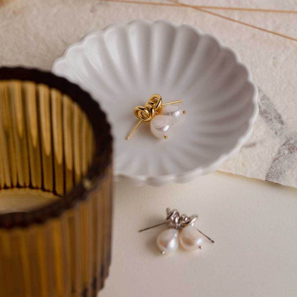 The Knot Pearl Drop Earrings in gold and silver are displayed on a white dish, set against a brown glass and textured background.