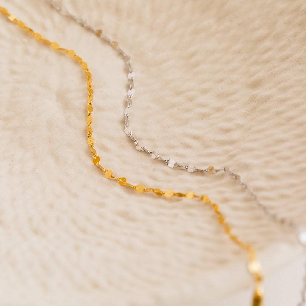 A gold chain and the Josephine Chain Bracelet are displayed on a textured, cream-colored surface.