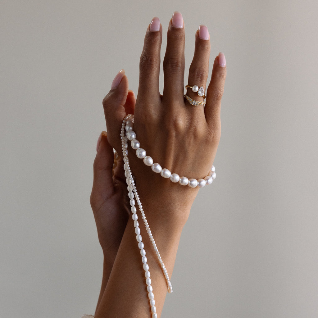 A hand with manicured nails holds a high-luster Pearl Choker Necklace and wears gold rings with pearls.