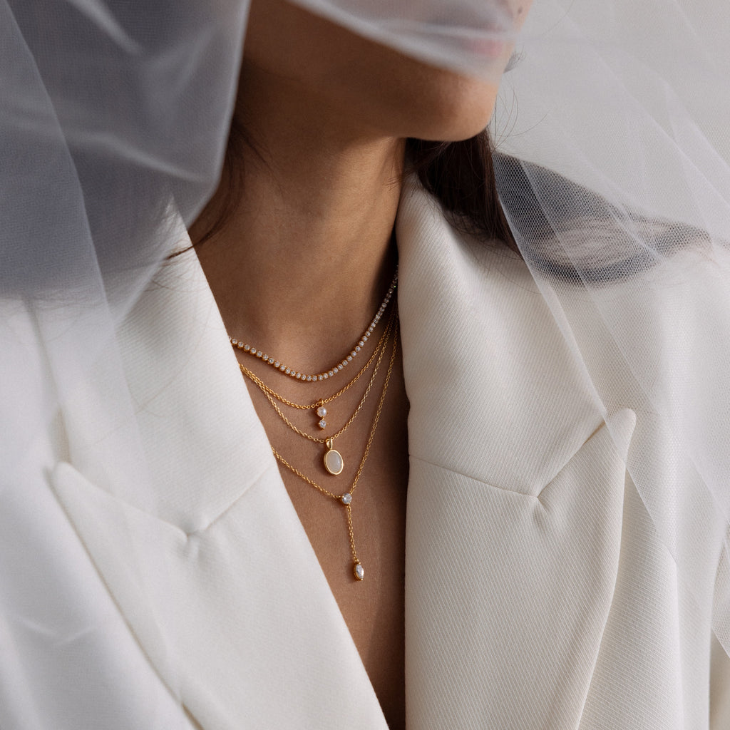 Woman in white blazer wears layered gold necklaces, featuring an Oval Labradorite Pendant Necklace, partially covered by a sheer white veil.