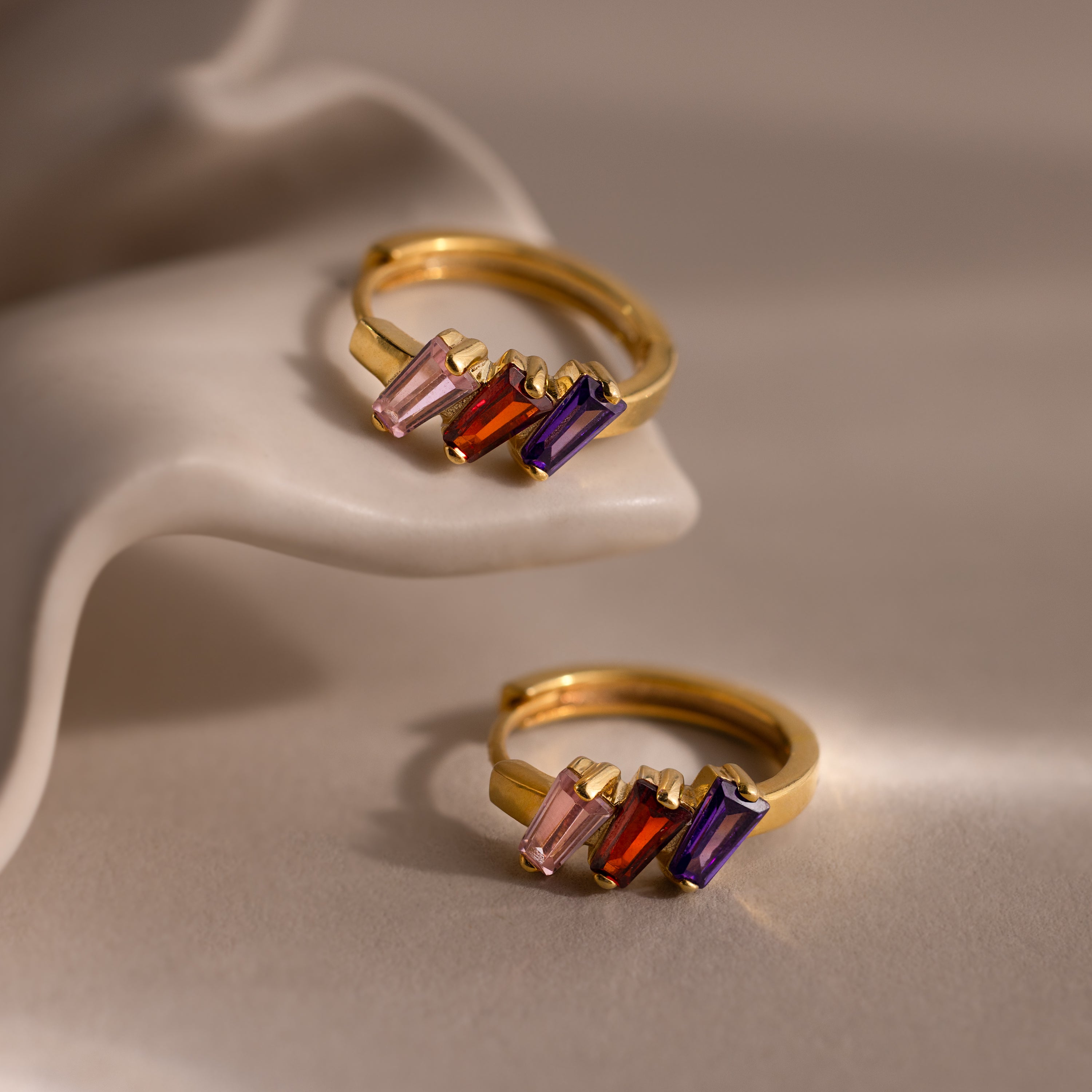 The Multiple Tapered Birthstone Huggies are two gold rings featuring pink, red, and purple rectangular stones, displayed on a curved beige surface—ideal for fans of modern statement jewelry.