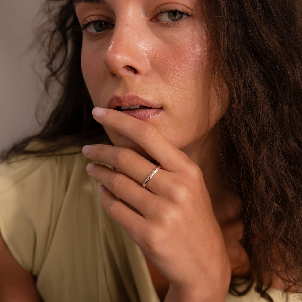 A woman with wavy brown hair touches her lips, wearing a cream top and the Diamond Morse Code Ring in Sterling Silver—a sleek band with a hidden message.