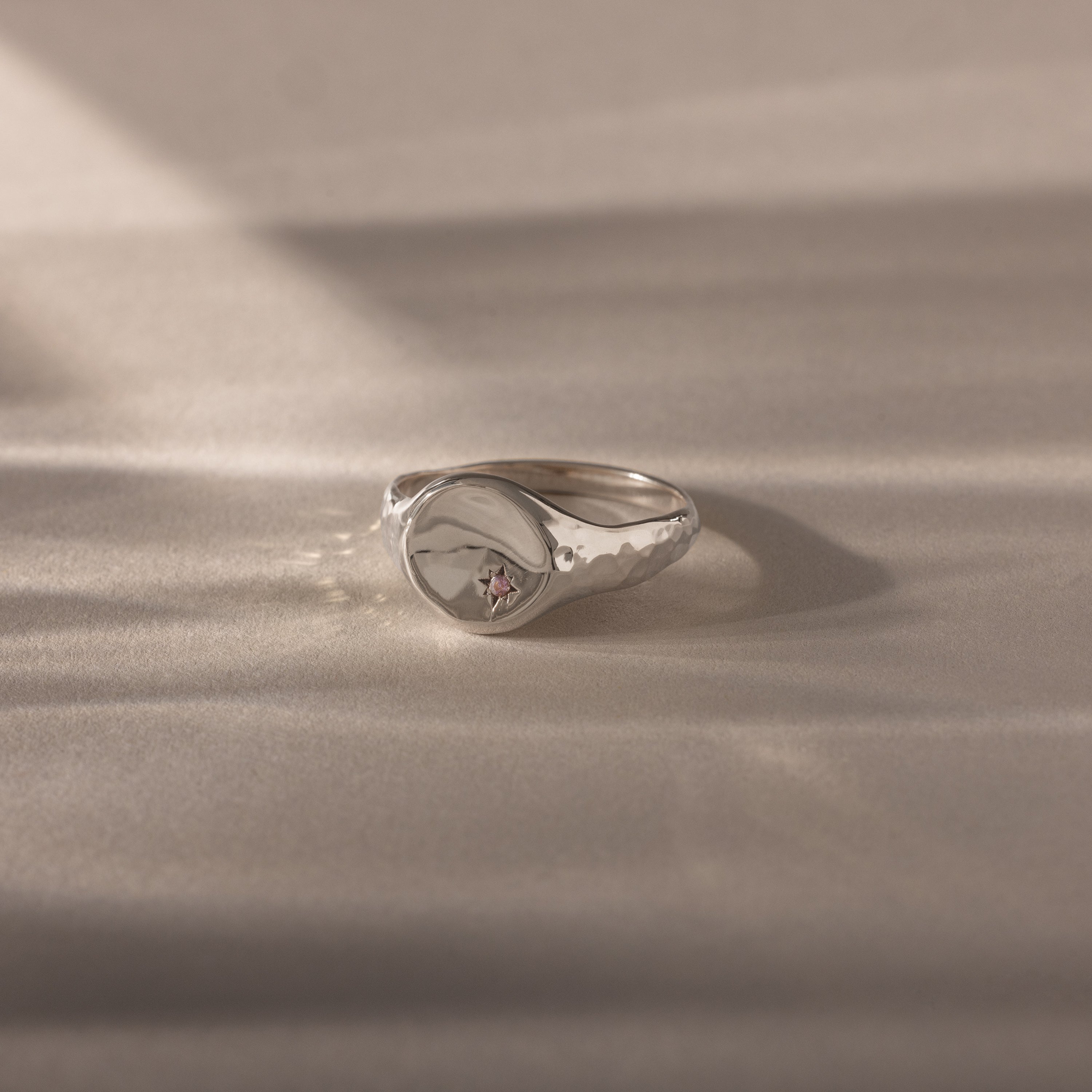 The North Star Alexandrite Signet Ring, featuring a star-shaped engraving with a pink alexandrite gemstone set in silver, rests on a soft beige surface—ideal for those who love meaningful jewelry.