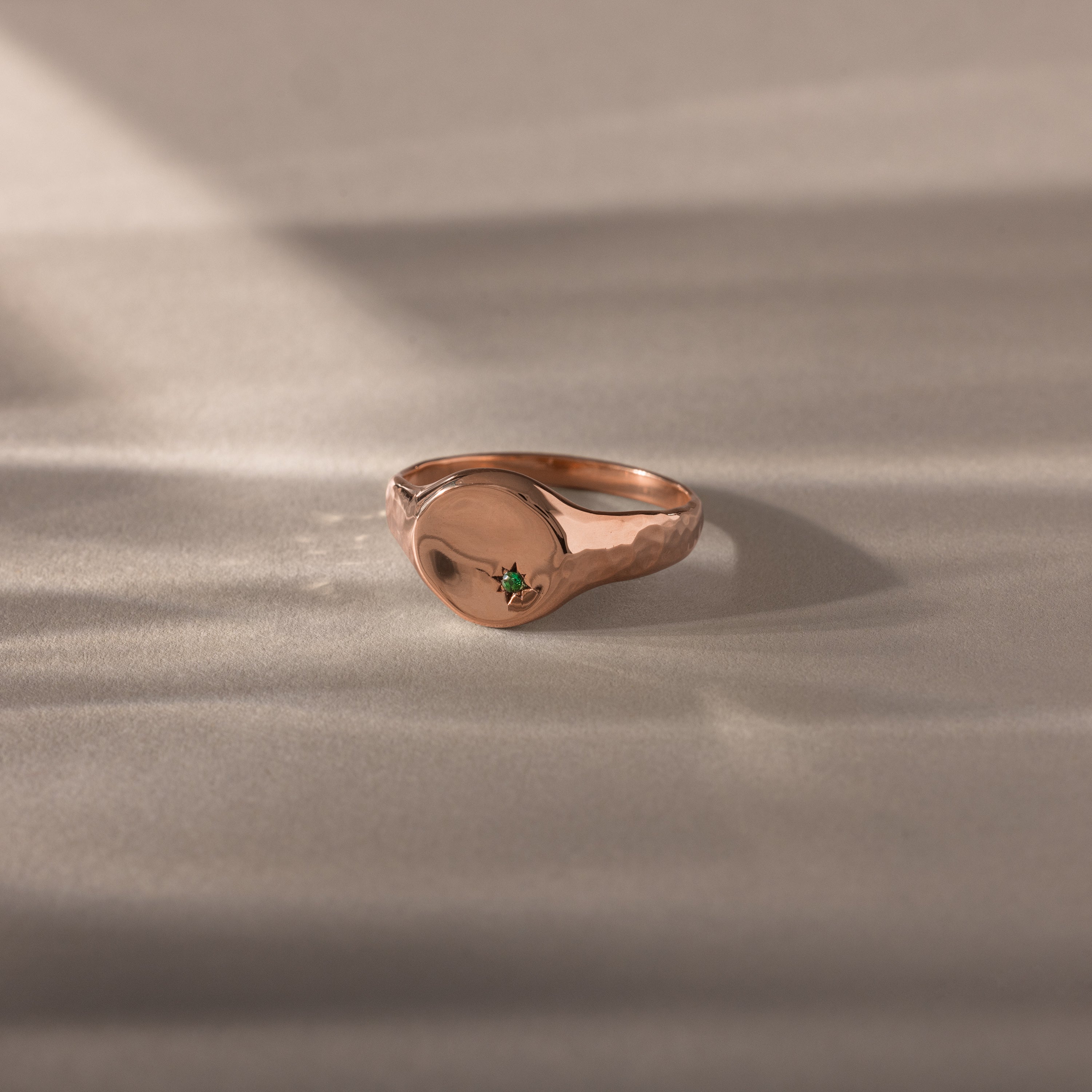 The North Star Emerald Signet Ring features a rose gold, smooth round face with a small green gemstone at the center, making it ideal for those seeking symbolic jewelry. Shown on a neutral surface.