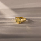 The North Star Birthstone Signet Ring in emerald (May) features a small green gemstone at its center and is showcased on a soft beige surface.
