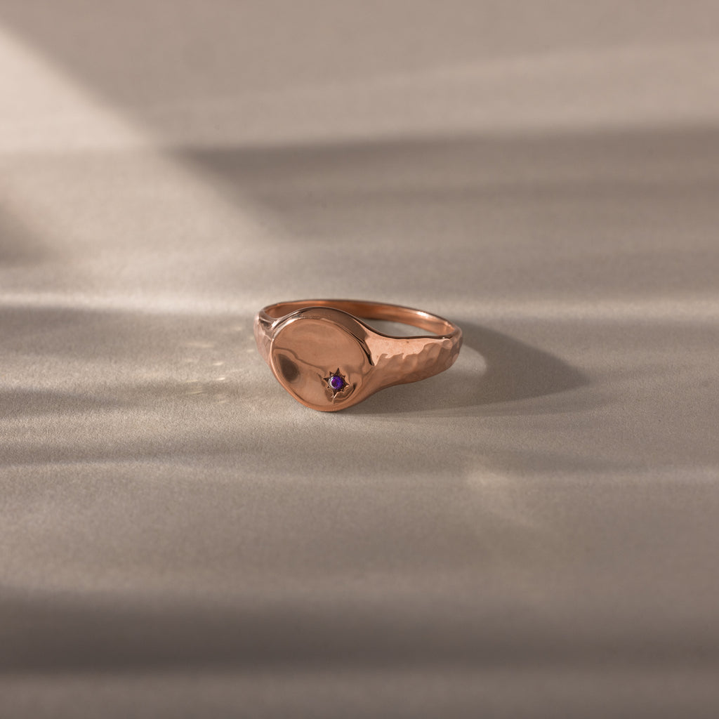 The North Star Amethyst Signet Ring in rose gold features a small purple gemstone, showcased on a light textured surface with soft shadows—an elegant piece of symbolic jewelry.