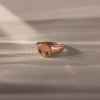 The North Star Amethyst Signet Ring in rose gold features a small purple gemstone, showcased on a light textured surface with soft shadows—an elegant piece of symbolic jewelry.