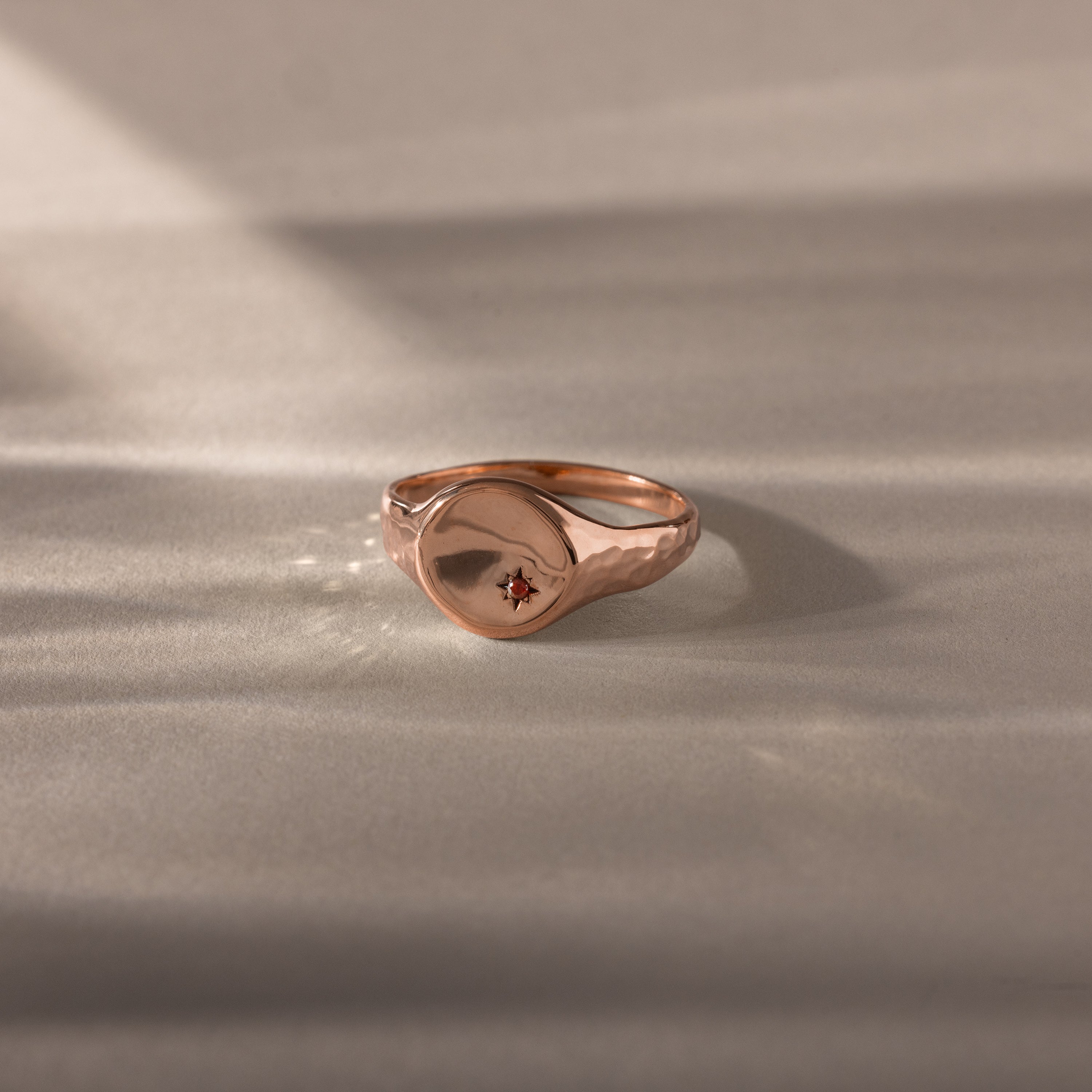 The North Star Garnet Signet Ring features a vintage-inspired gold design with a small red garnet centerpiece, elegantly displayed on a neutral surface—an exquisite piece of symbolic jewelry.