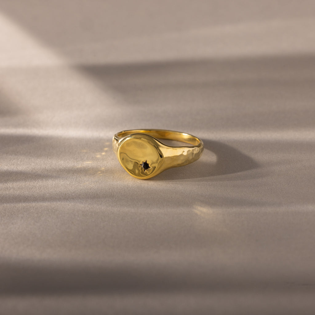 The North Star Birthstone Signet Ring, featuring an onyx center stone, is a symbolic gold ring displayed elegantly on a light, textured surface.