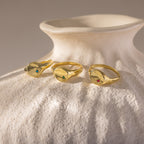 Three gold rings, including the North Star Birthstone Signet Ring with colorful gemstones, rest on a textured white ceramic vase in soft light.