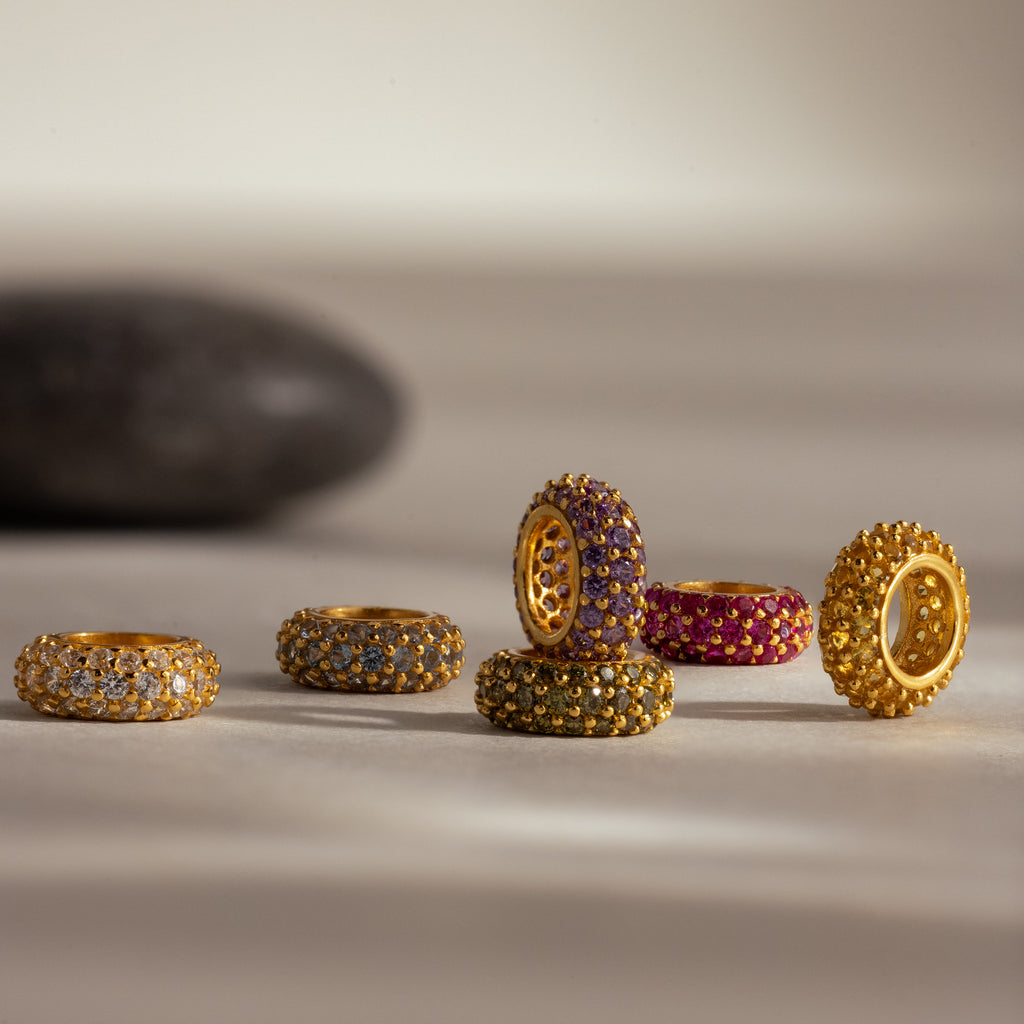 Six gold rings with colorful gemstones, including a Pave Topaz Charm Spacer, are arranged on a light surface with a dark stone in the background for added elegance to this jewelry display.