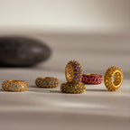 Six gold rings with colorful gemstones, including a Pave Topaz Charm Spacer, are arranged on a light surface with a dark stone in the background for added elegance to this jewelry display.