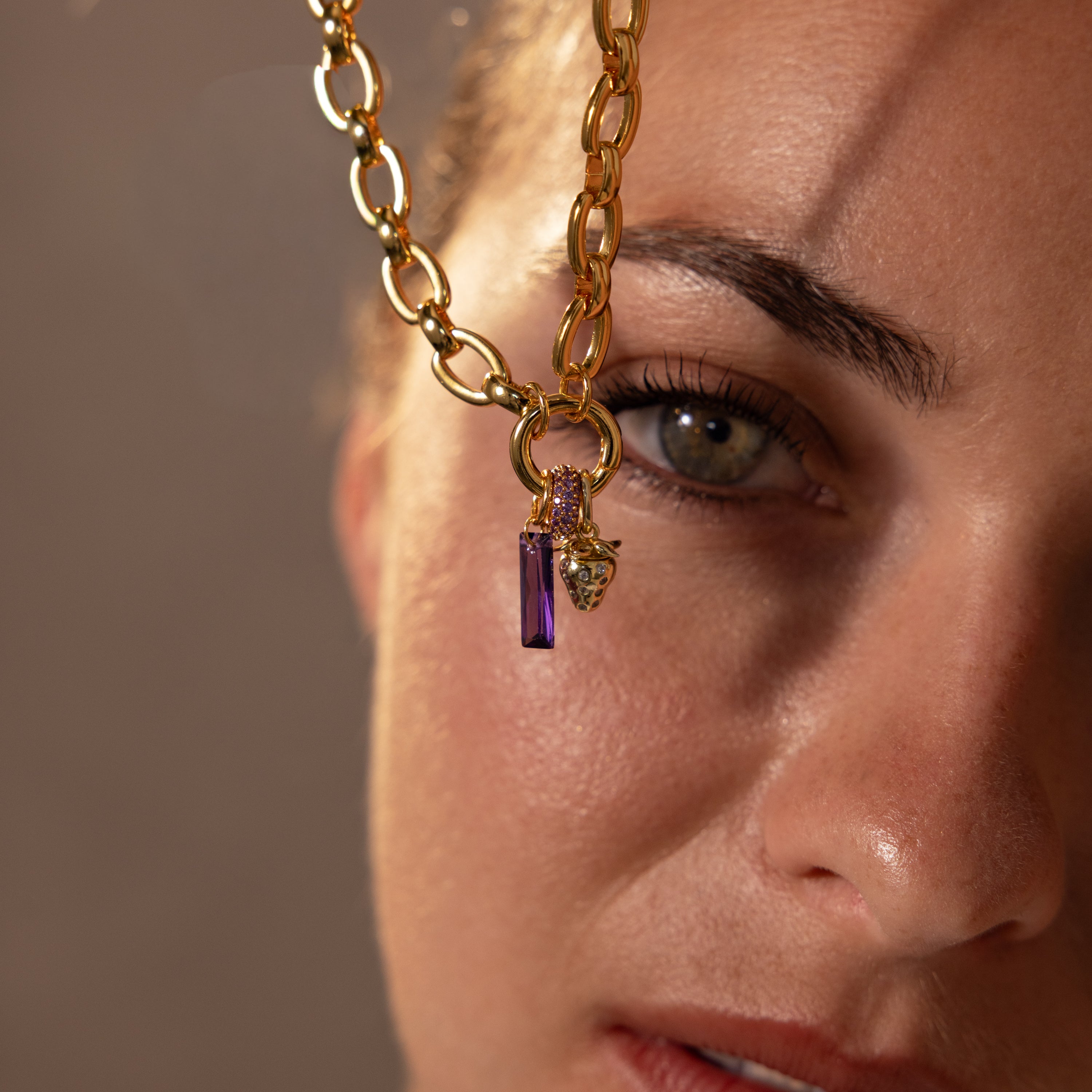 A close-up of a womans face wearing a gold chain necklace with the Pave Birthstone Charm Spacer, showcasing its purple pendant in front of her eye.