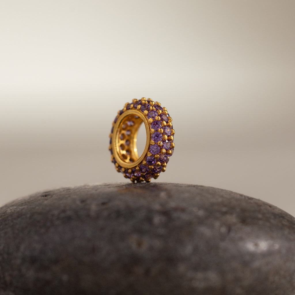 The Pave Birthstone Charm Spacer, a gold circular piece with purple gemstones, sits upright on a smooth, dark stone surface.