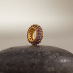 The Pave Birthstone Charm Spacer, a gold circular piece with purple gemstones, sits upright on a smooth, dark stone surface.