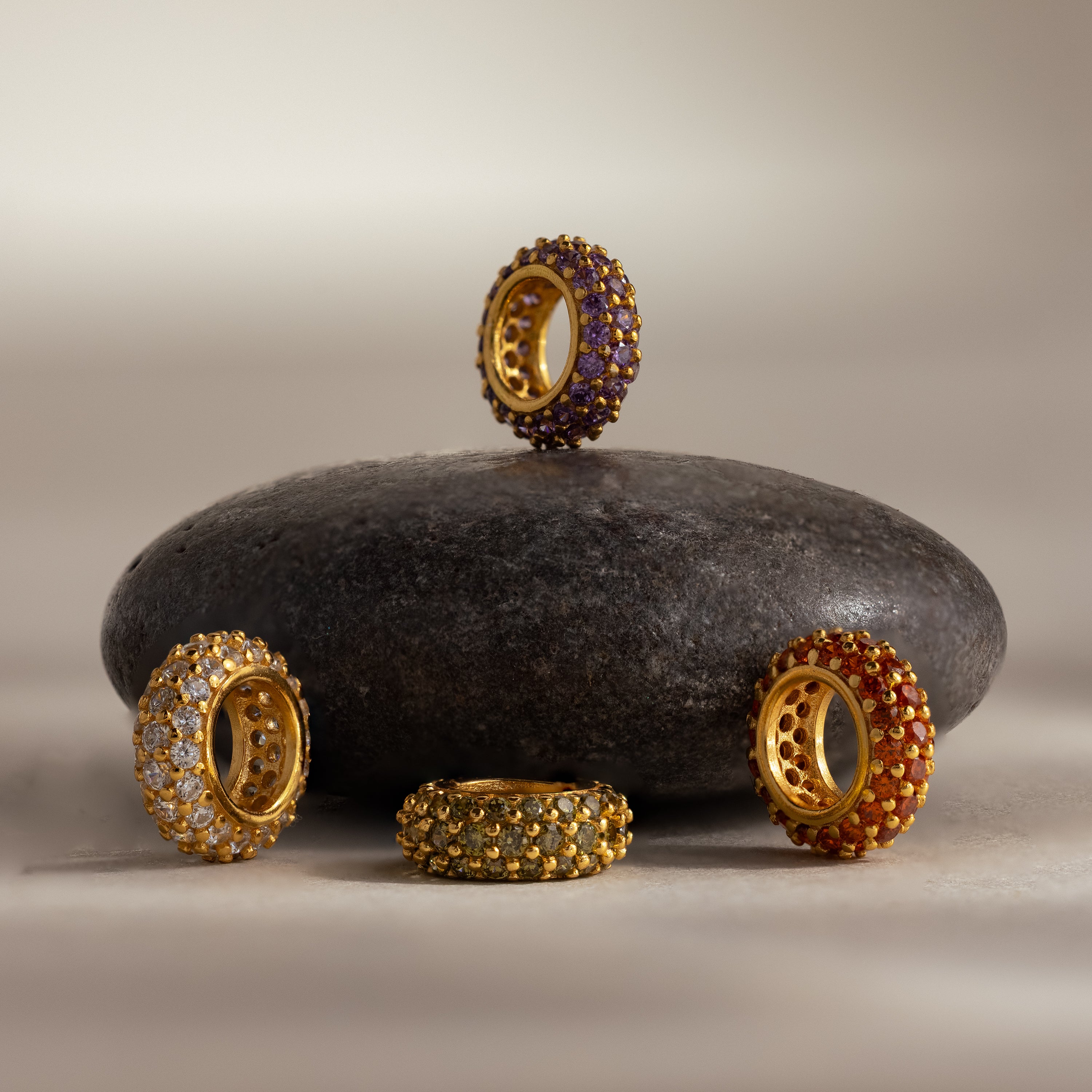 Five ornate gold rings with colorful gemstones encircle a smooth, dark stone, capturing the elegance of birthstone jewelry and the beauty of the Pavé Birthstone Charm Spacer.