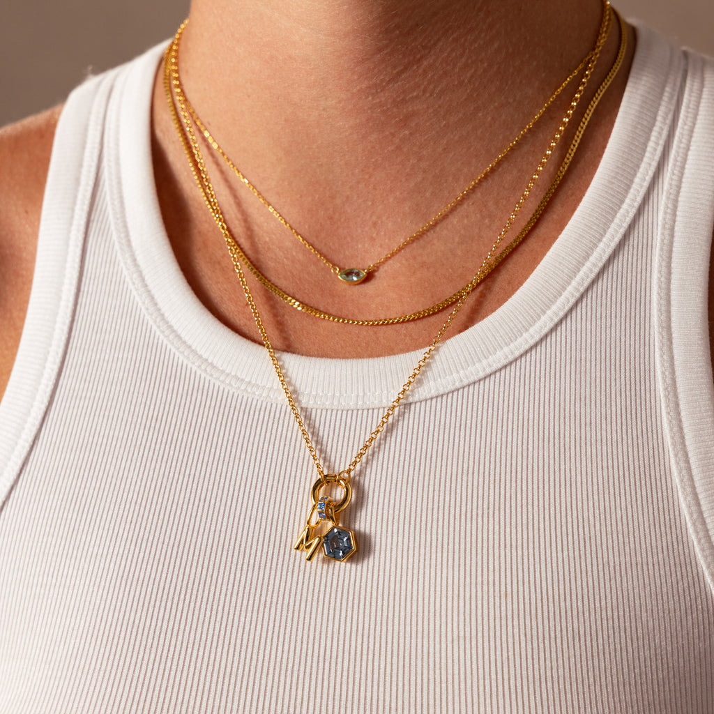 Close-up of a person wearing layered gold necklaces, featuring the Hexagon Birthstone Charm with a geometric frame, paired with a white ribbed tank—an ideal meaningful gift.