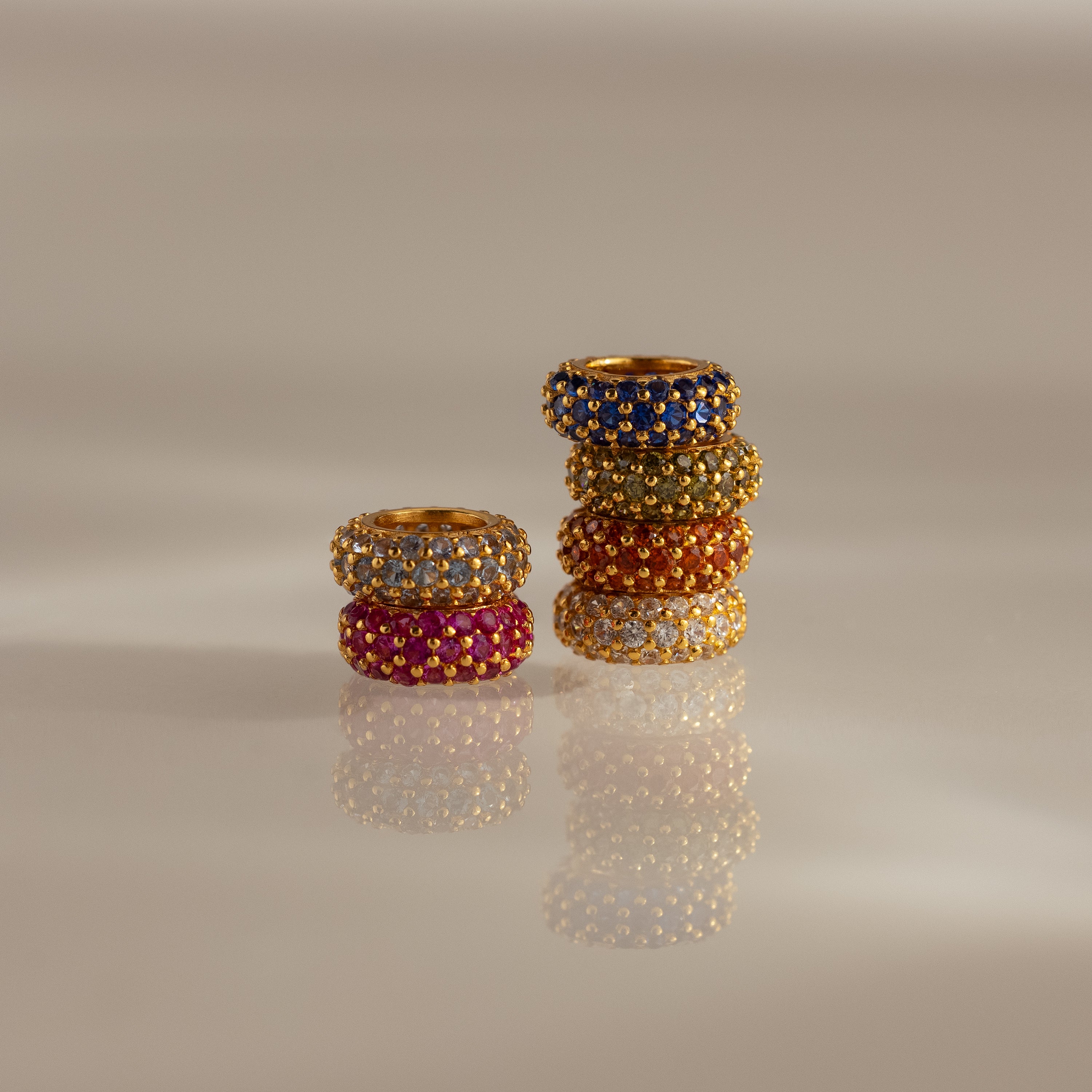 Six Pave Birthstone Charm Spacer rings with colorful gemstones are stacked in two groups on a glossy, beige surface.