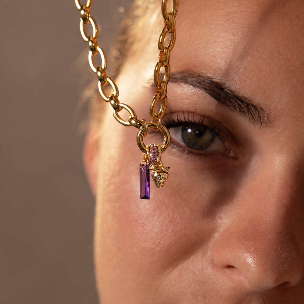 A close-up of a womans face with gold jewelry—a chain necklace featuring the Skinny Baguette Sapphire Charm—held near her eye.