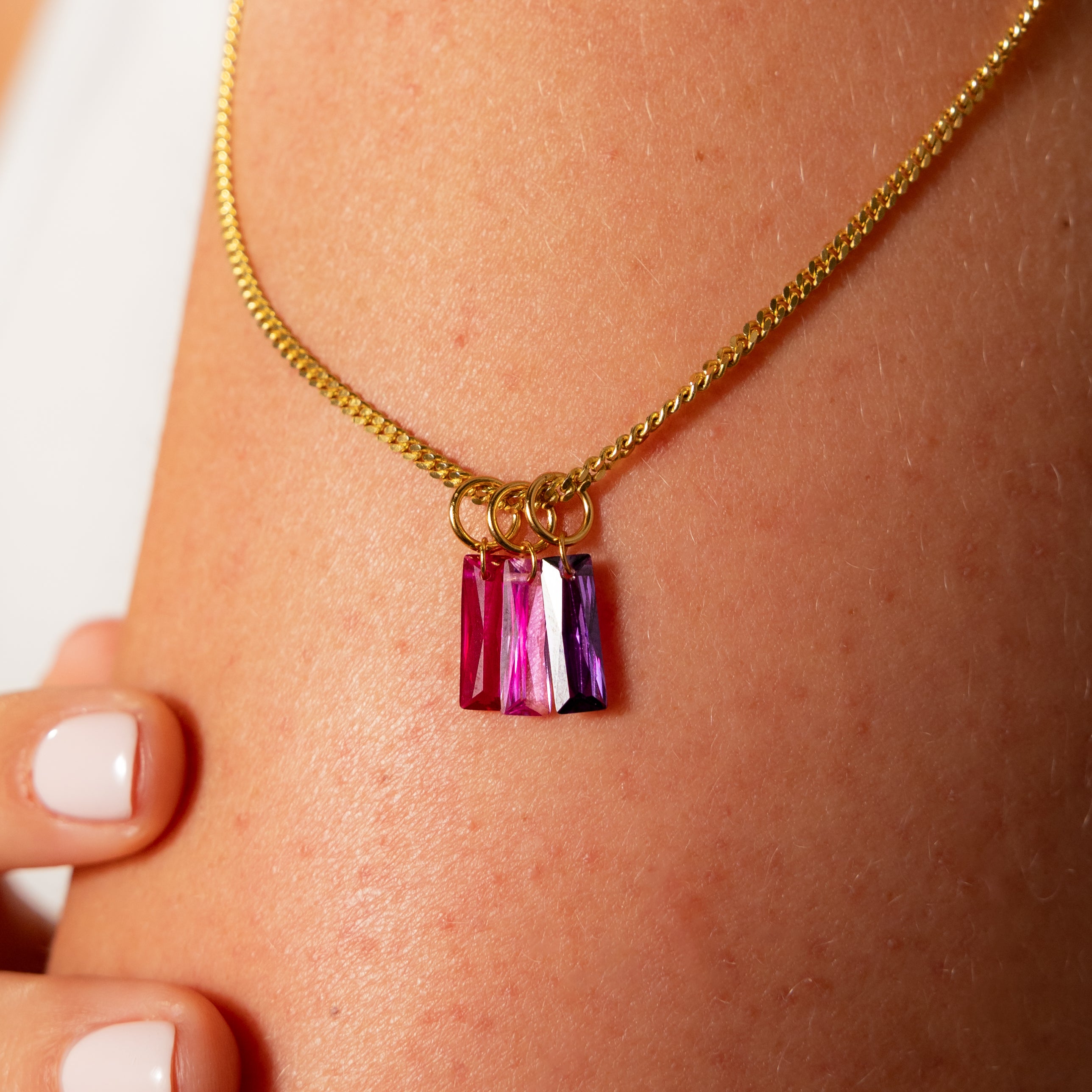The Skinny Baguette Sapphire Charm—a gold necklace with three baguette-cut pink and purple crystal pendants—rests elegantly on a persons neck.