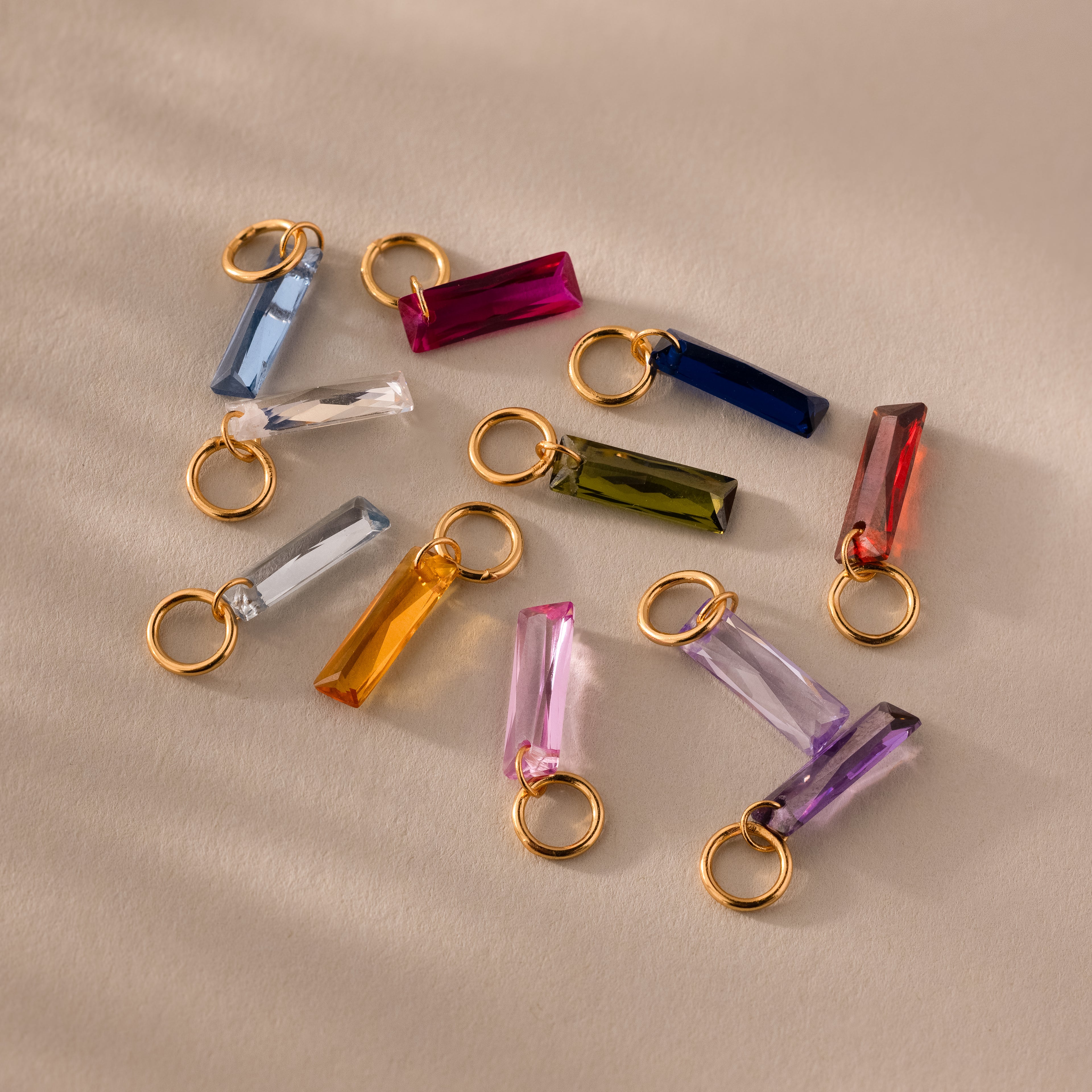 The Skinny Baguette Sapphire Charm features a colorful crystal baguette with a gold loop, ideal as a birthstone charm or an accent for gold jewelry, shown arranged on a beige surface.