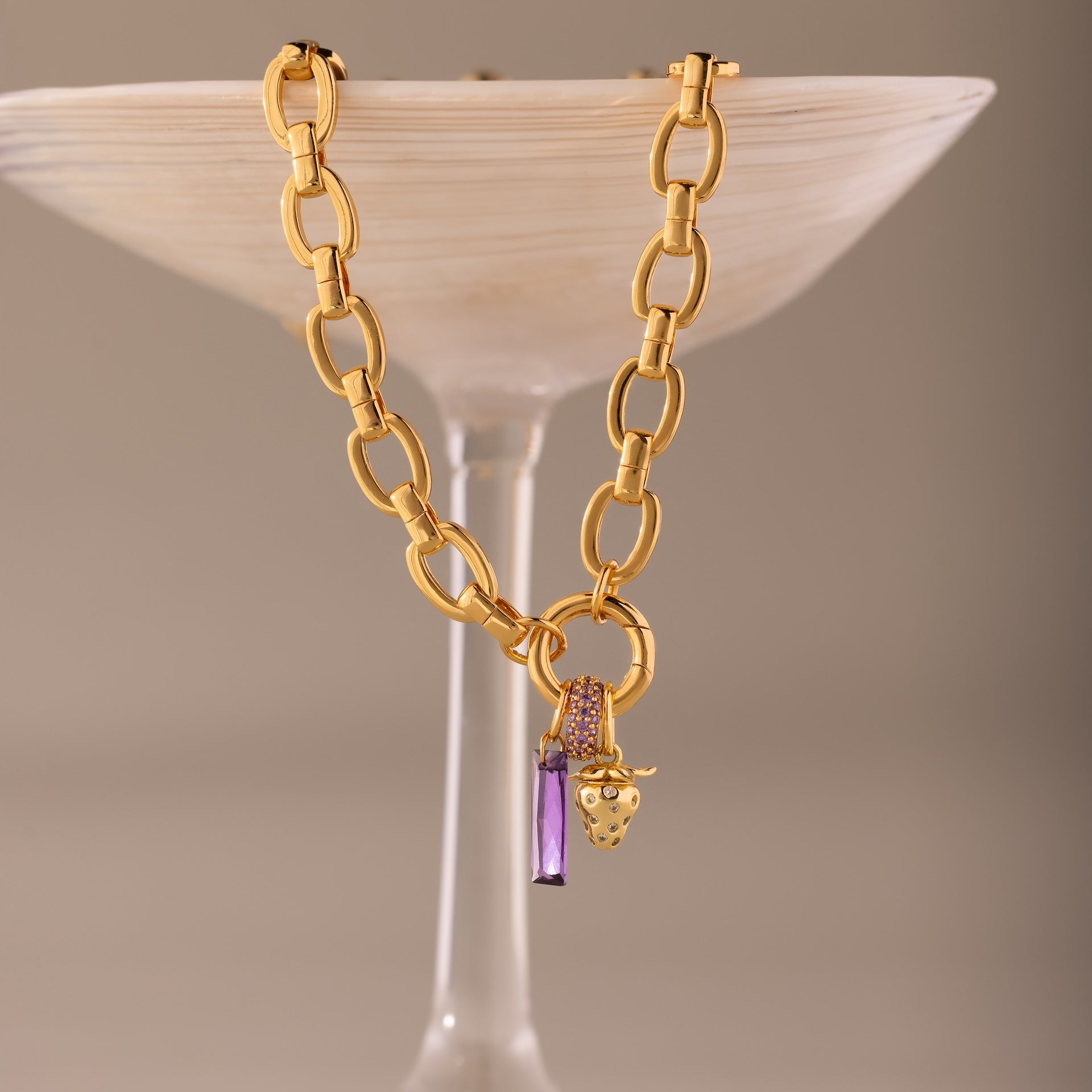 The Skinny Baguette Sapphire Charm, a gold chain necklace with sapphire and gold grape charms, drapes over frosted glass, featuring a sparkling birthstone accent for added elegance.