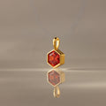 The Hexagon Garnet Charm is a gold pendant with a faceted hexagonal red gem, showcased on a smooth neutral surface—an eye-catching piece of geometric jewelry.