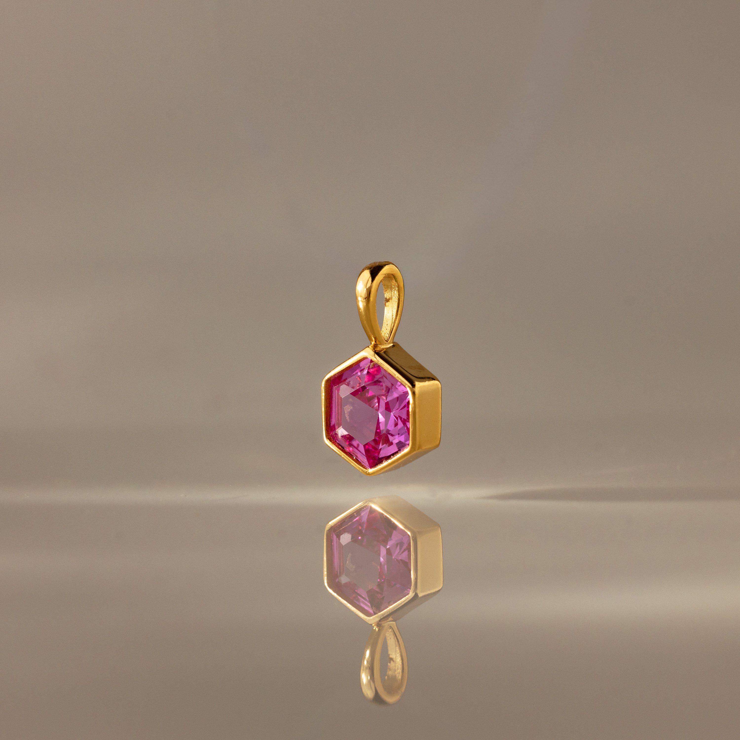 The Hexagon Pink Tourmaline Charm features a gold hexagonal pendant with a faceted pink stone, elegantly displayed on a smooth, neutral surface—a modern geometric jewelry piece.