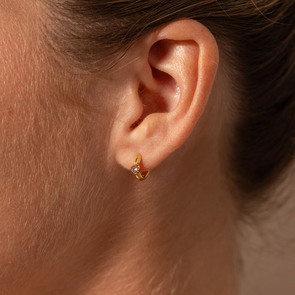 Close-up of a person’s ear wearing Ribbed Emerald Huggies—small gold hoop earrings with a single emerald gemstone. A perfect, meaningful gift for any occasion.