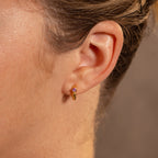 Close-up of a person’s ear wearing Ribbed Ruby Huggies gold hoop earrings, accented with a pink ruby gemstone for a subtle touch of birthstone jewelry.