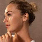 A blonde-haired woman in a bun wears Ribbed Birthstone Huggies and gazes thoughtfully leftward against a neutral background.