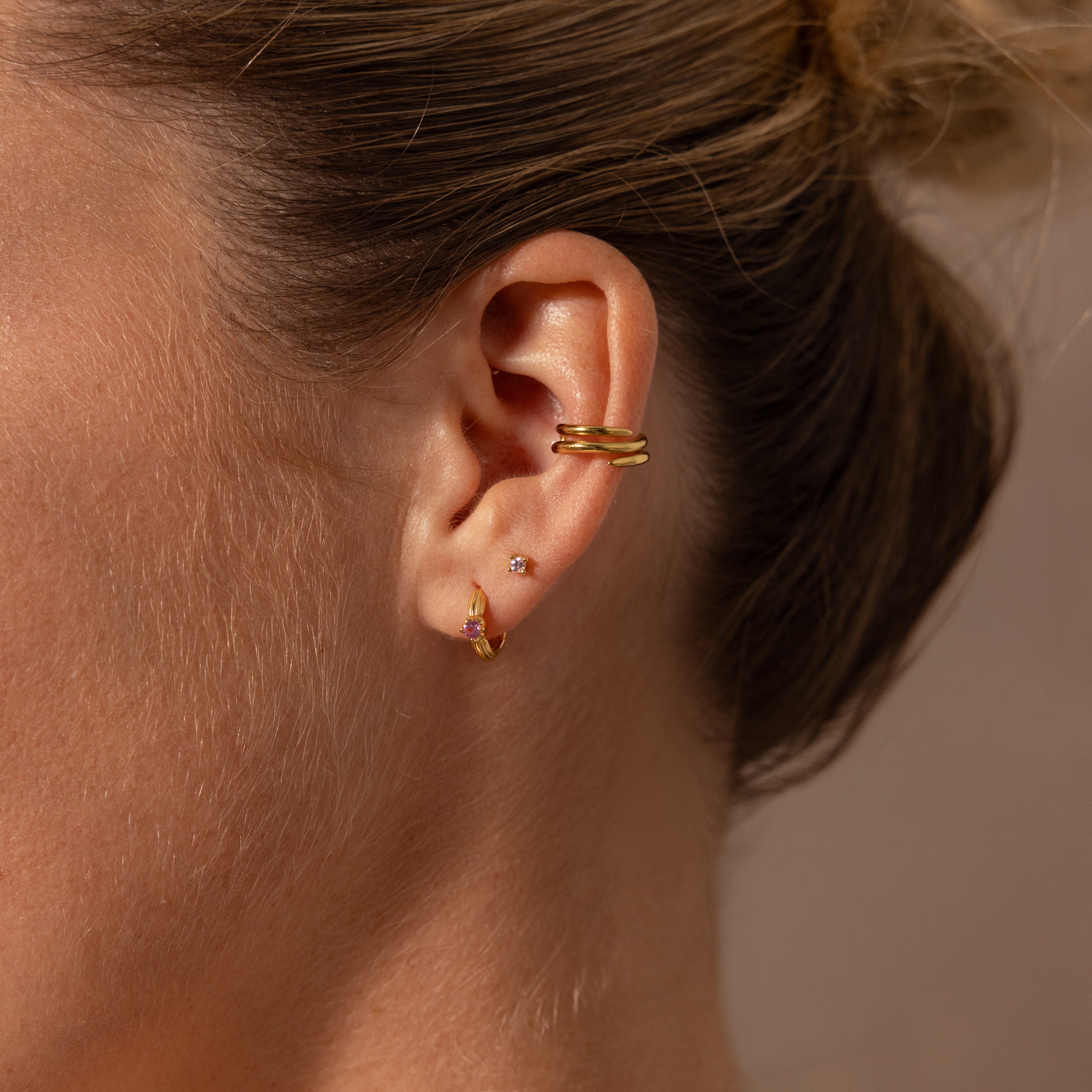 Close-up of a womans ear with three gold earrings: a double cuff, a small stud, and Ribbed Birthstone Huggies—gold hoops featuring a pink birthstone.