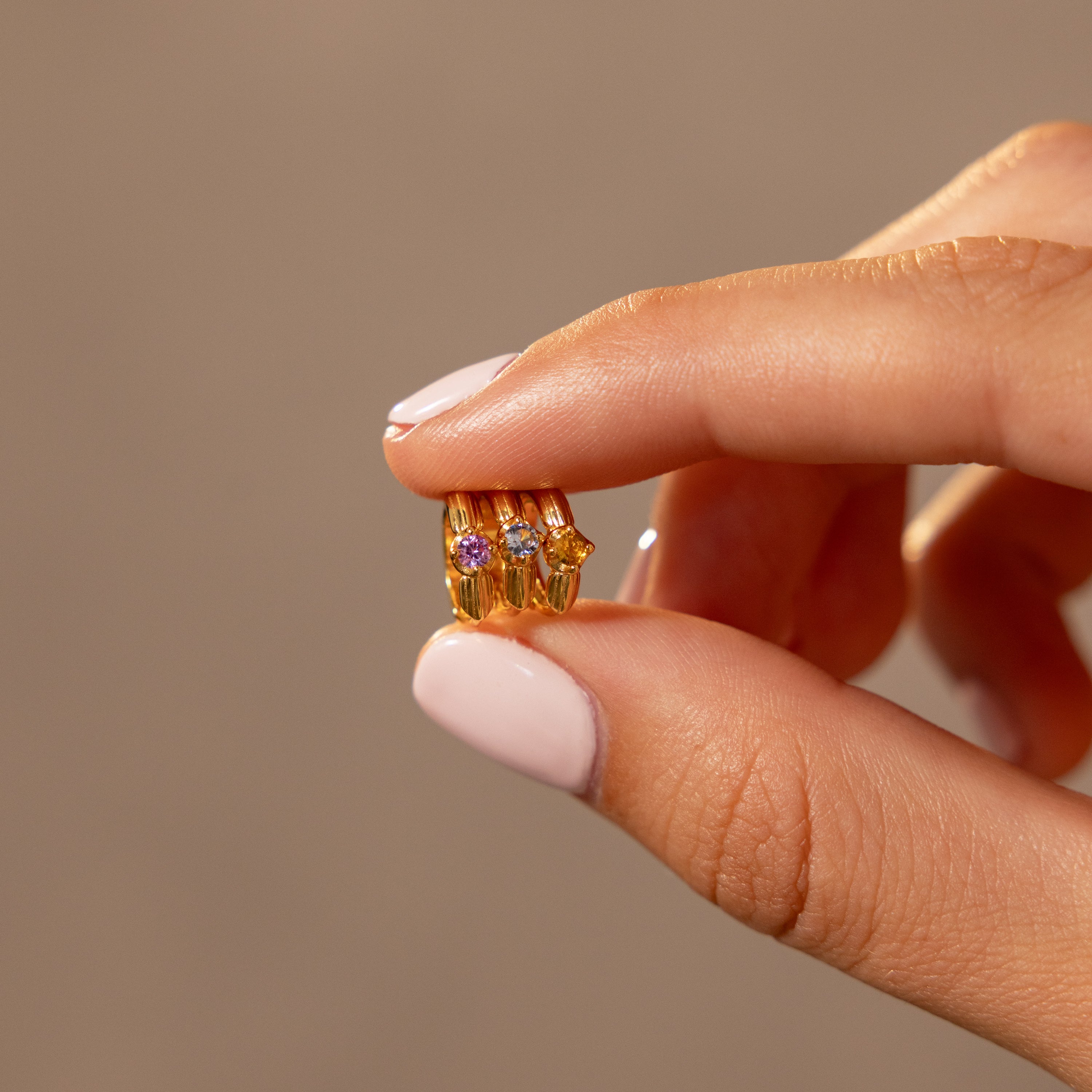 A hand with pale pink nails holds a small gold ring, similar to the Ribbed Birthstone Huggies, decorated with three colorful gemstones.