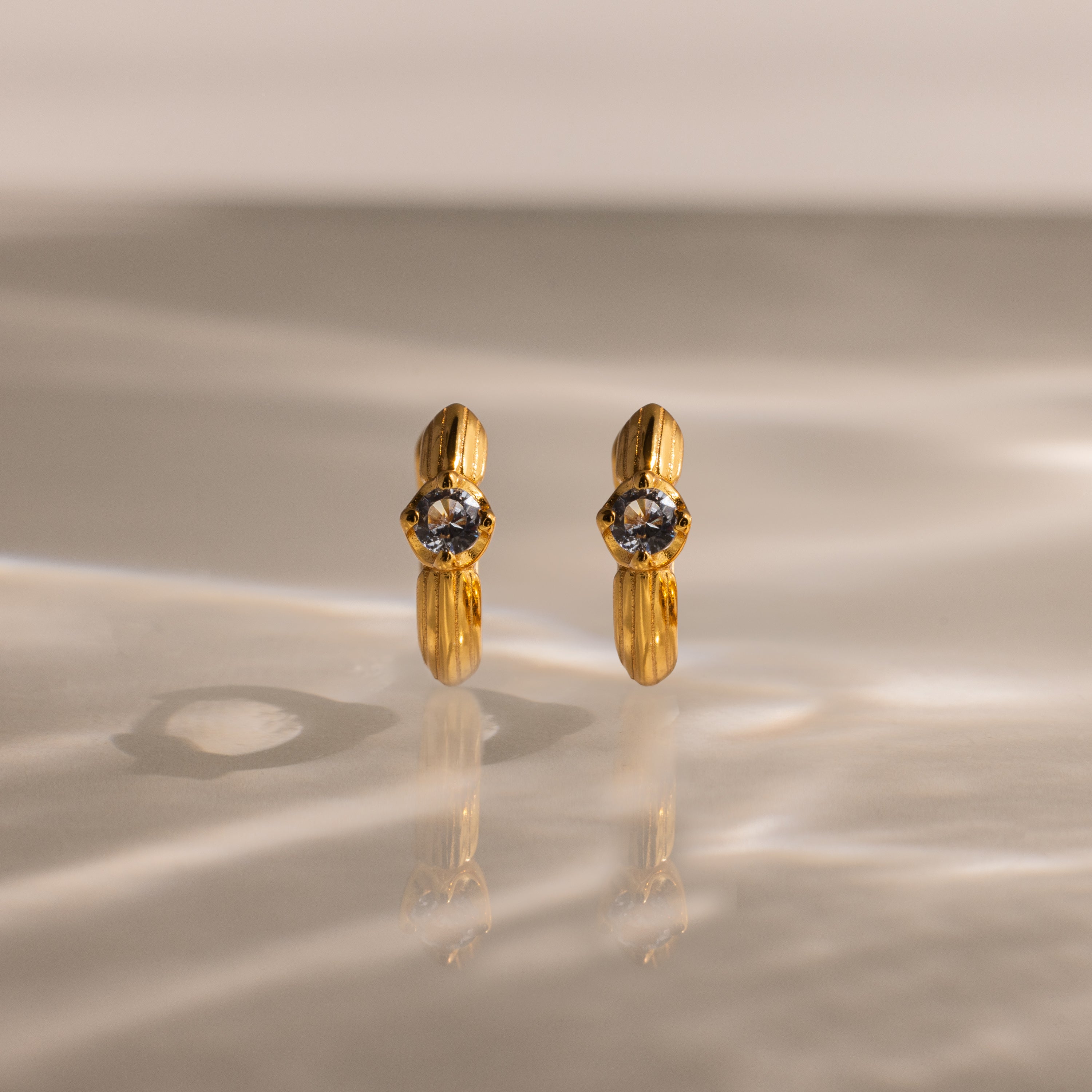Gold hoop earrings with clear gemstones rest on a reflective surface—these Ribbed Aquamarine Huggies bring personalized elegance to your jewelry collection.