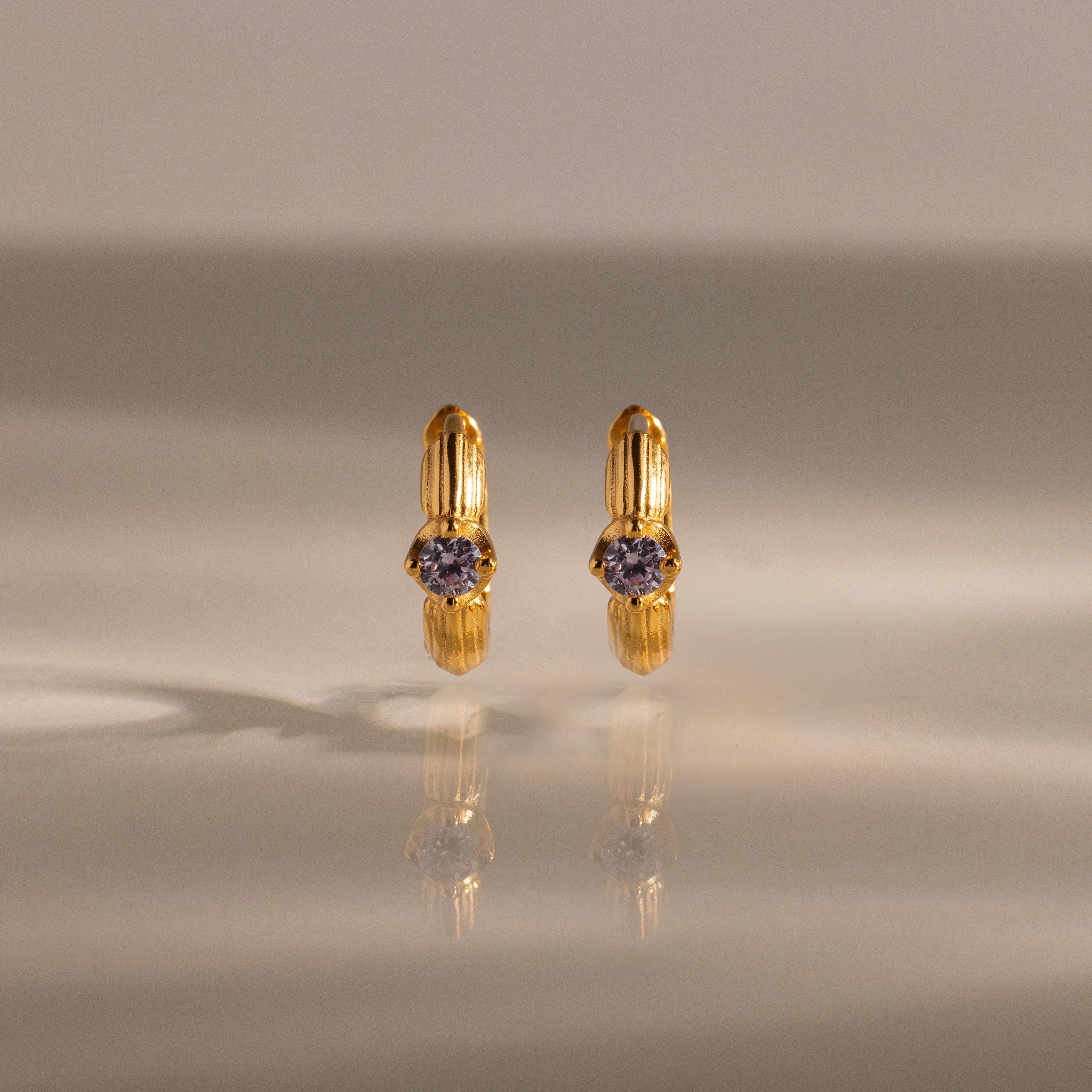 Ribbed Alexandrite Huggies, gold hoop earrings with a small round purple gemstone, stand upright on a reflective surface.