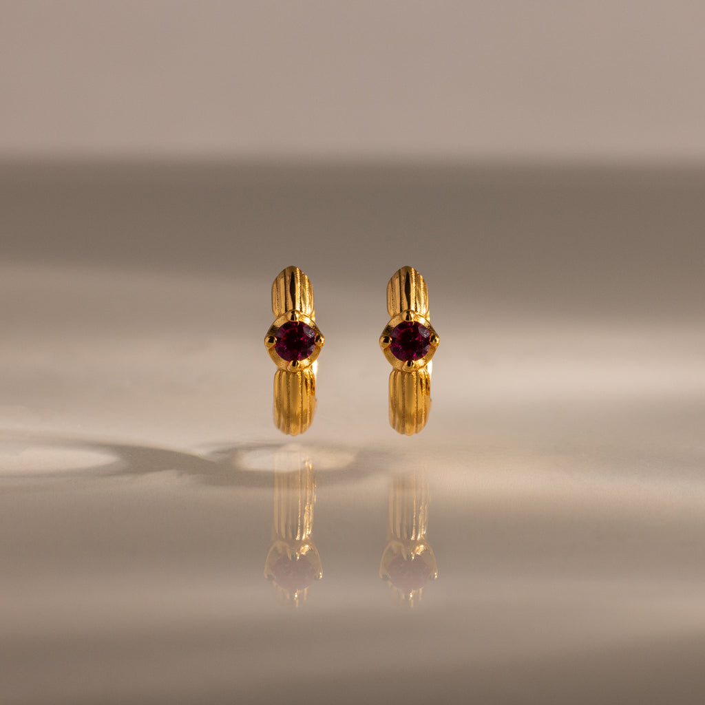 A pair of Ribbed Ruby Huggies with red gemstones stands upright on a reflective surface, illuminated by soft lighting.