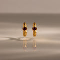 A pair of Ribbed Ruby Huggies with red gemstones stands upright on a reflective surface, illuminated by soft lighting.