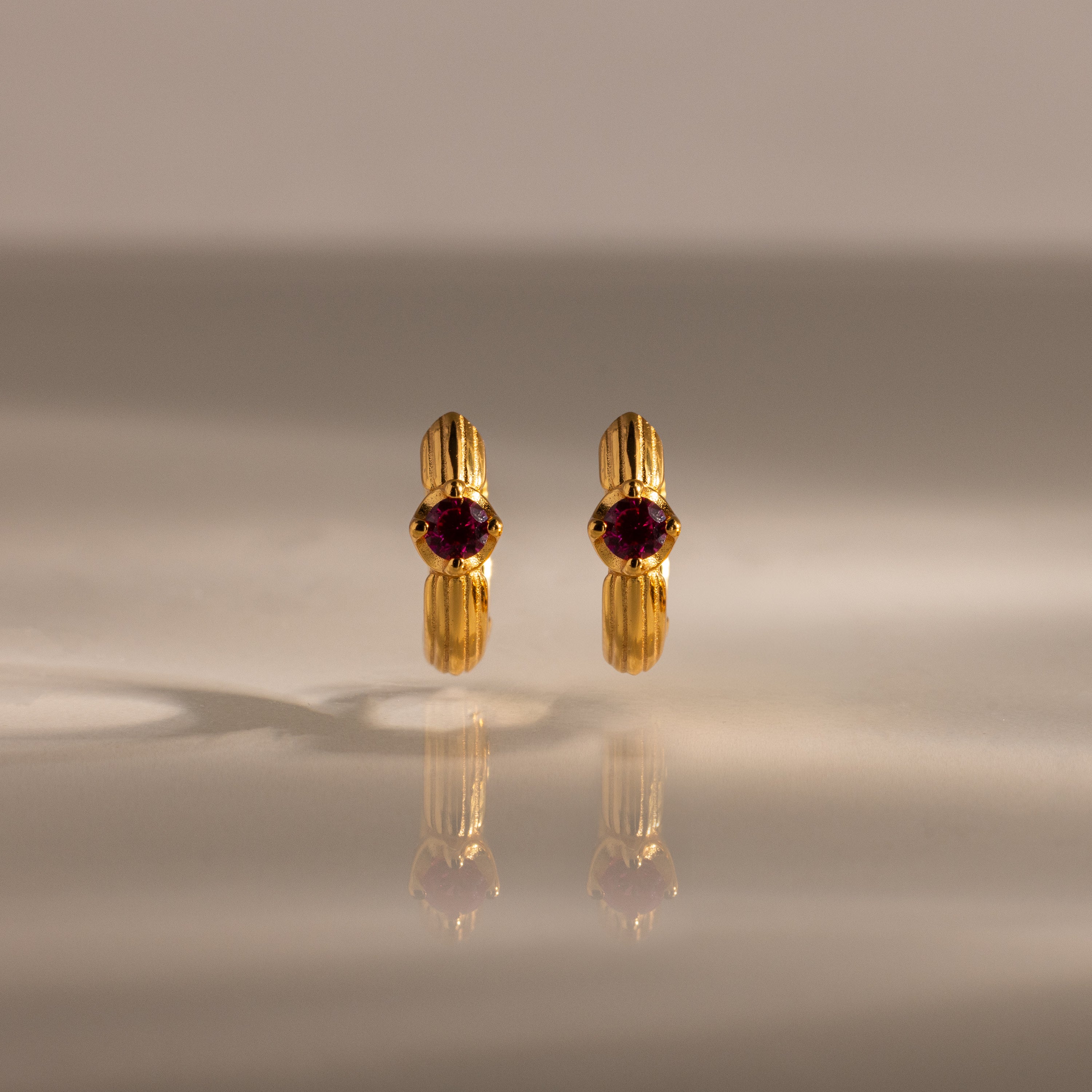 A pair of Ribbed Ruby Huggies with red gemstones stands upright on a reflective surface, illuminated by soft lighting.