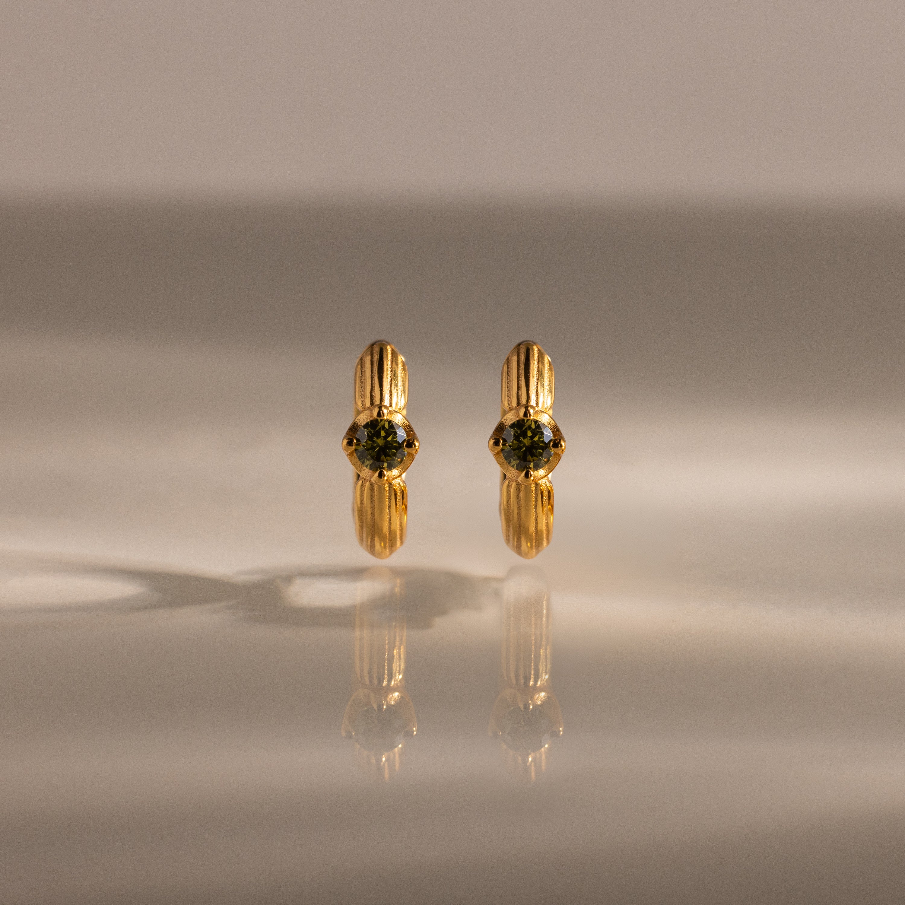 Ribbed Peridot Huggies—gold stud earrings with round green gemstones, shown on a reflective beige surface with soft lighting—ideal for lovers of personalized jewelry.