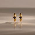 Ribbed Sapphire Huggies—gold stud earrings with blue gemstones—displayed upright on a reflective surface with a soft background, make a meaningful gift for any occasion.