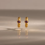 A pair of Ribbed Pink Tourmaline Huggies with pink gemstones, standing upright on a reflective surface—gold hoop earrings that make a meaningful gift.
