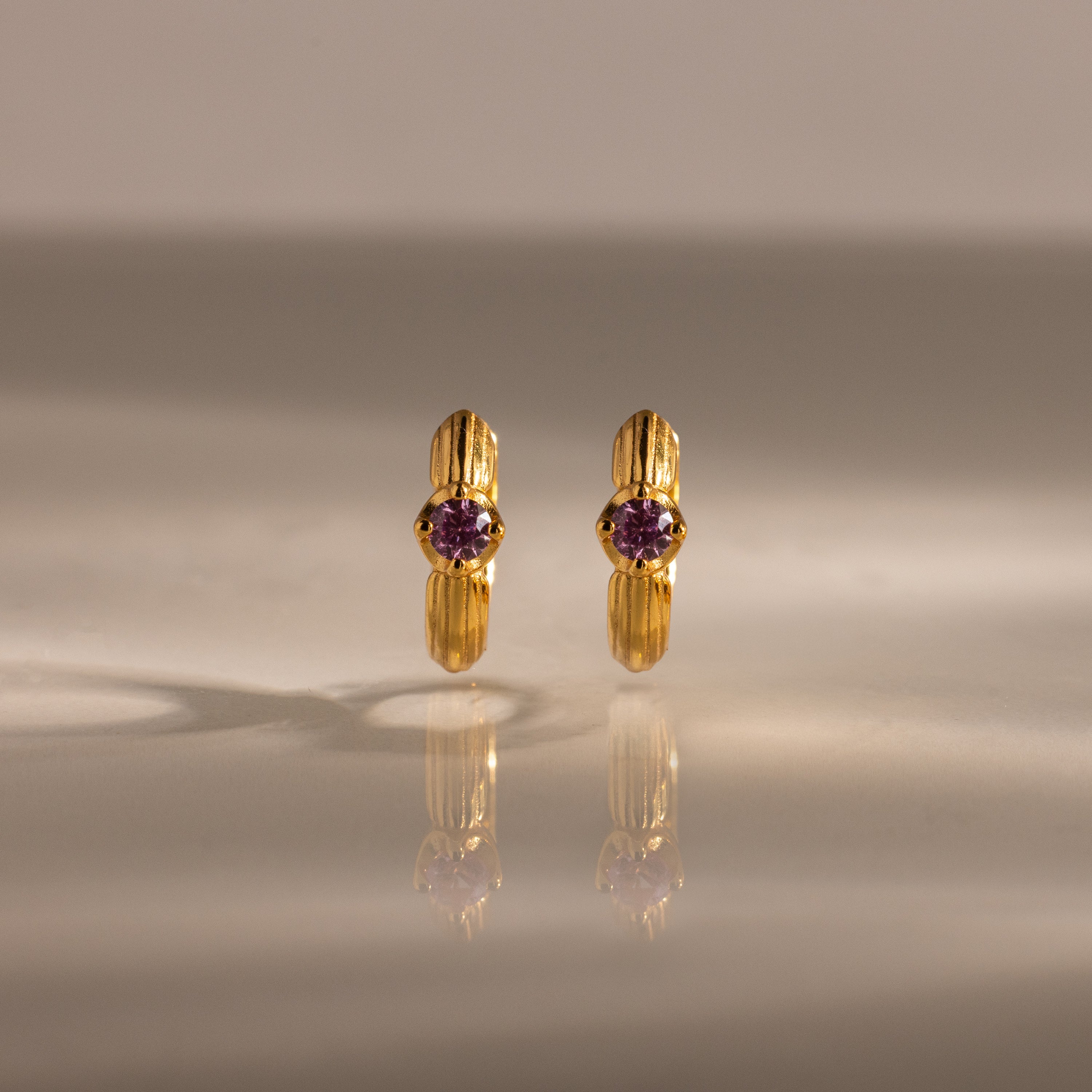A pair of Ribbed Pink Tourmaline Huggies with pink gemstones, standing upright on a reflective surface—gold hoop earrings that make a meaningful gift.