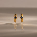 The Ribbed Blue Zircon Huggies, gold hoop earrings with blue zircon centers, are shown on a reflective surface, making this personalized jewelry sparkle.