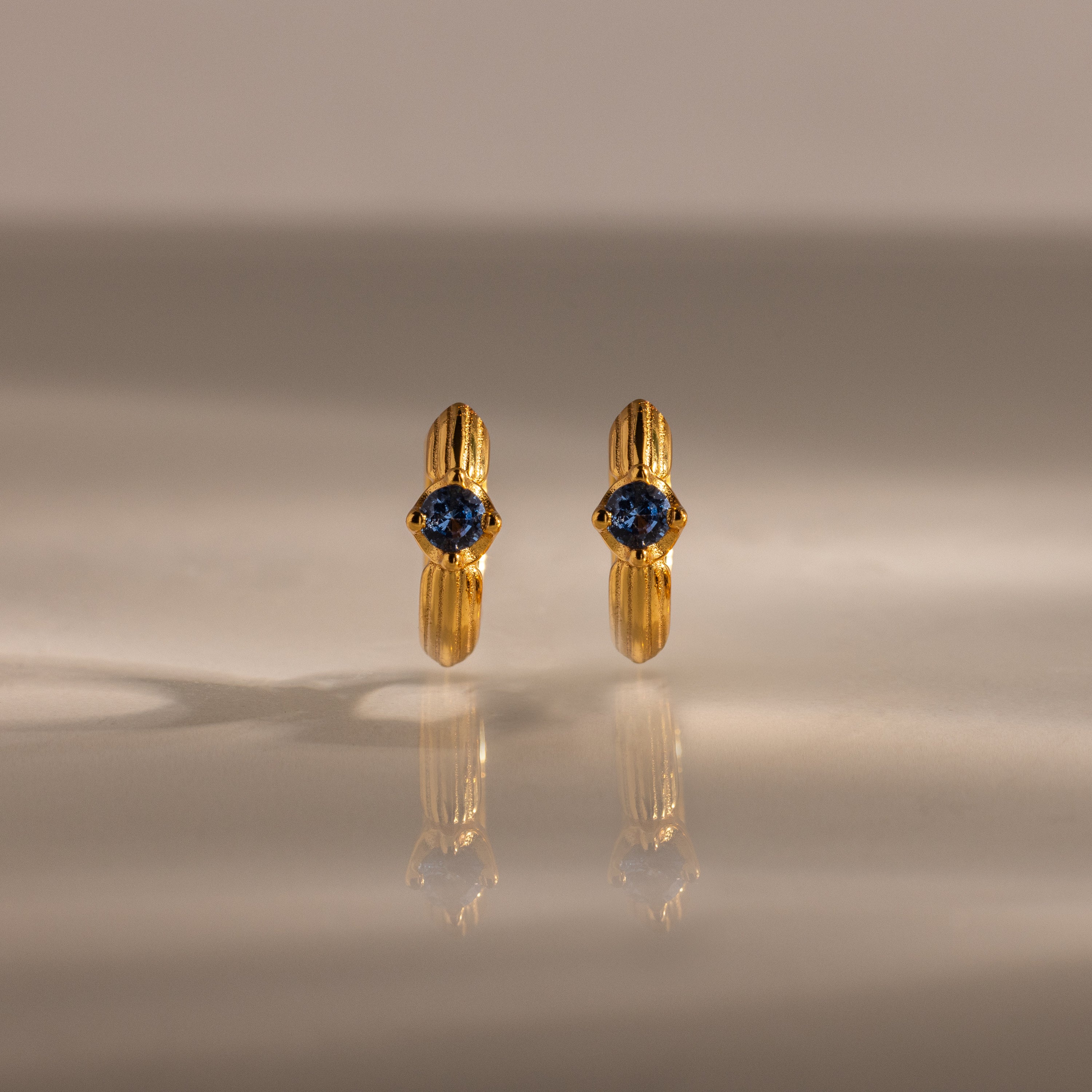 The Ribbed Blue Zircon Huggies, gold hoop earrings with blue zircon centers, are shown on a reflective surface, making this personalized jewelry sparkle.